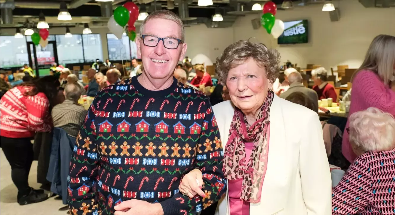One man and one woman standing arm in arm in front of a festive decorated background.