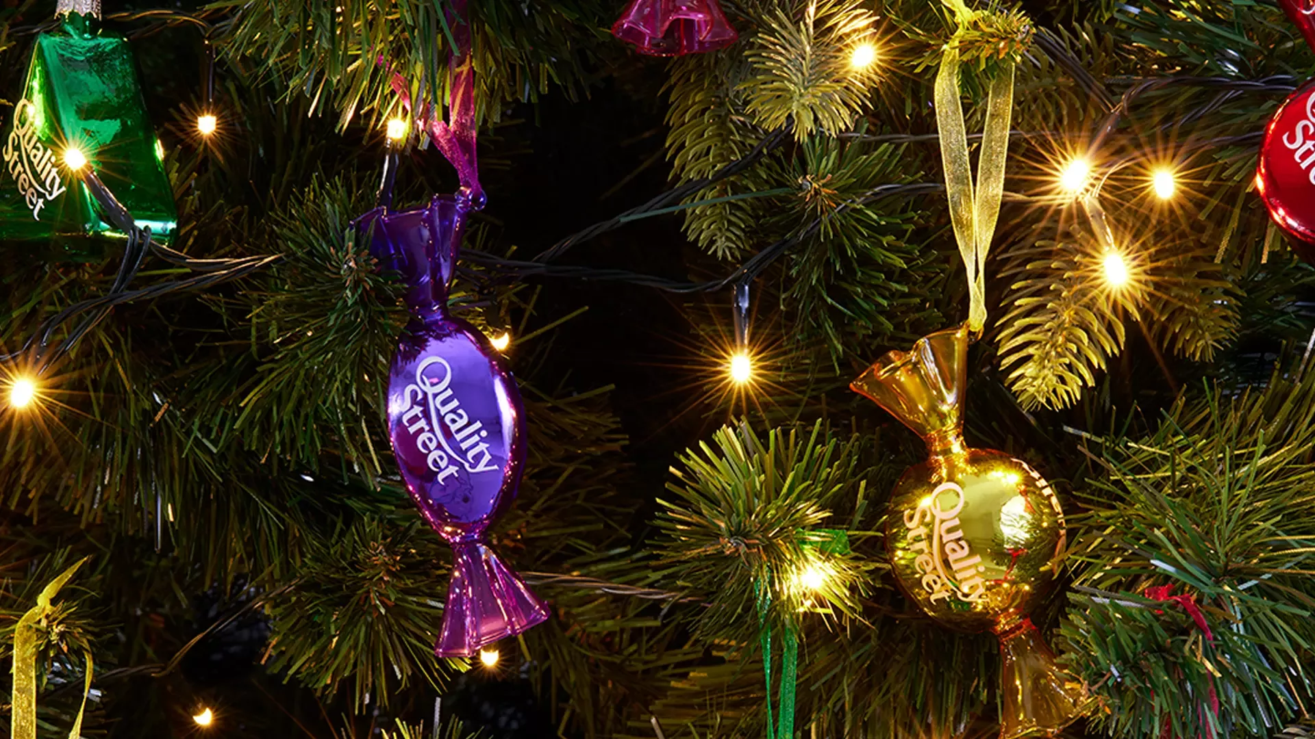 A close up photo of two purple and gold Quality Street Christmas baubles, on a green Christmas tree. 