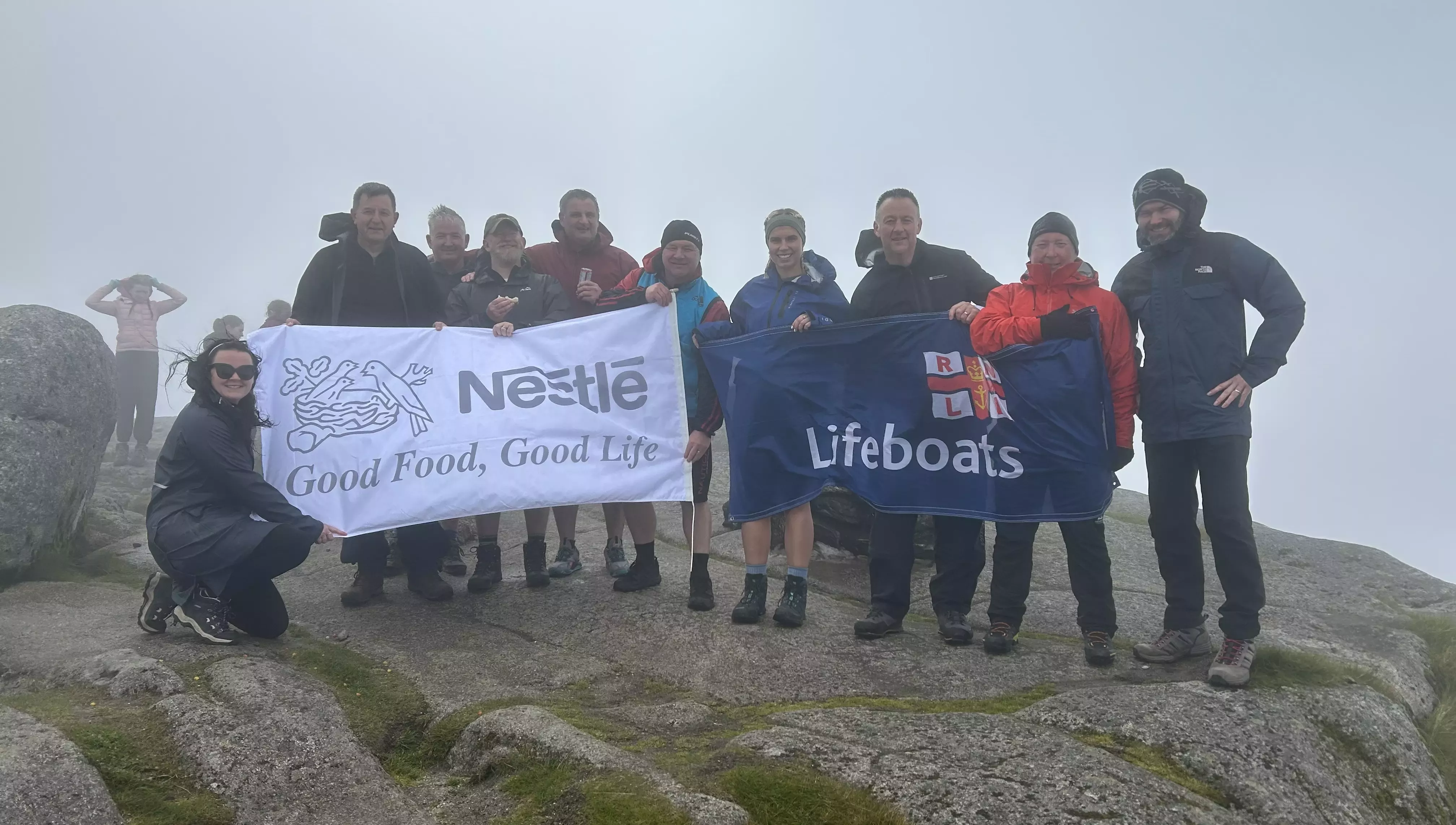 A group of ten people standing on top of a misty mountain with a Nestlé banner and a RNLI banner. 