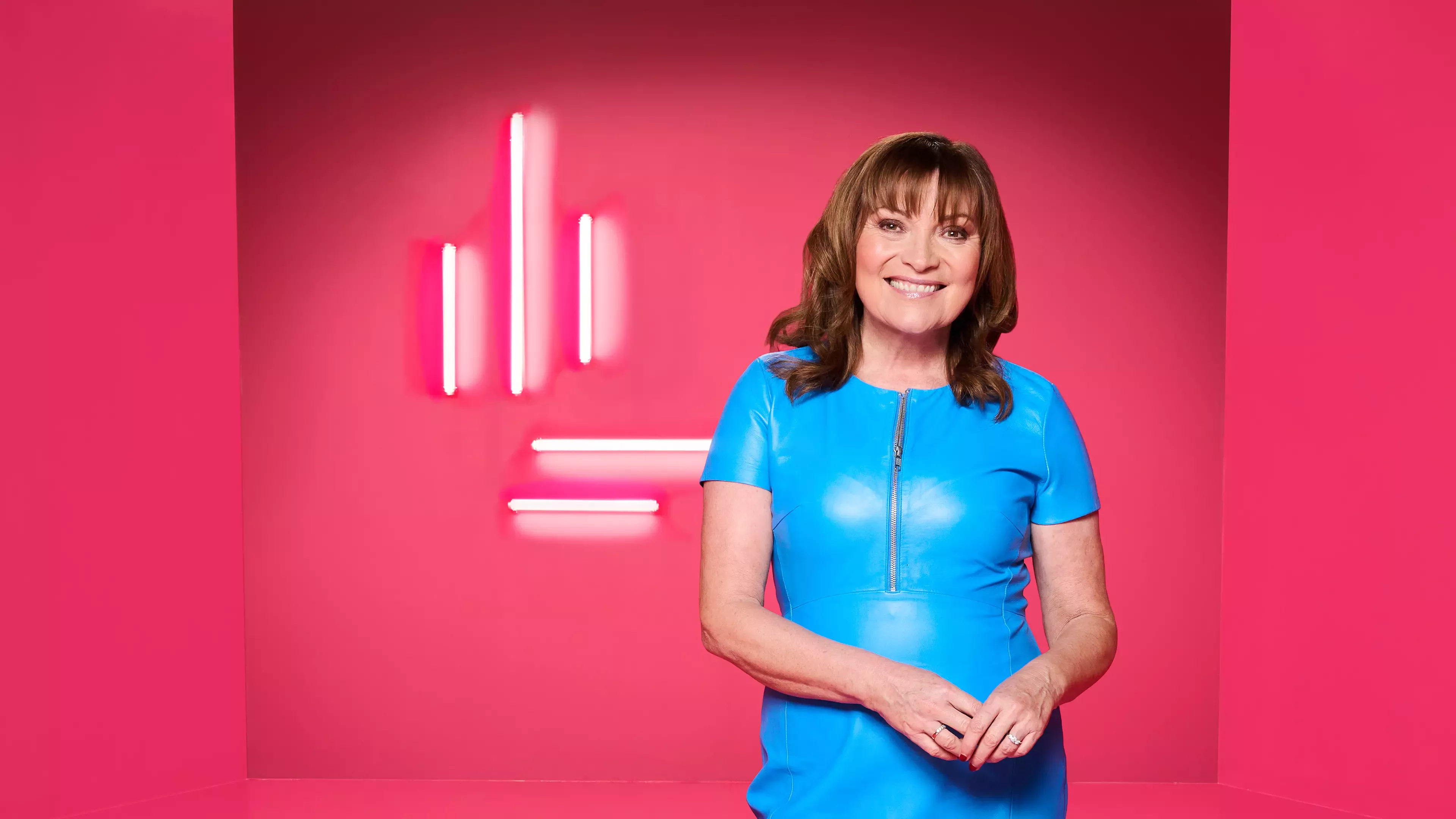 Lady with brown hair and blue dress in front of a pink background.