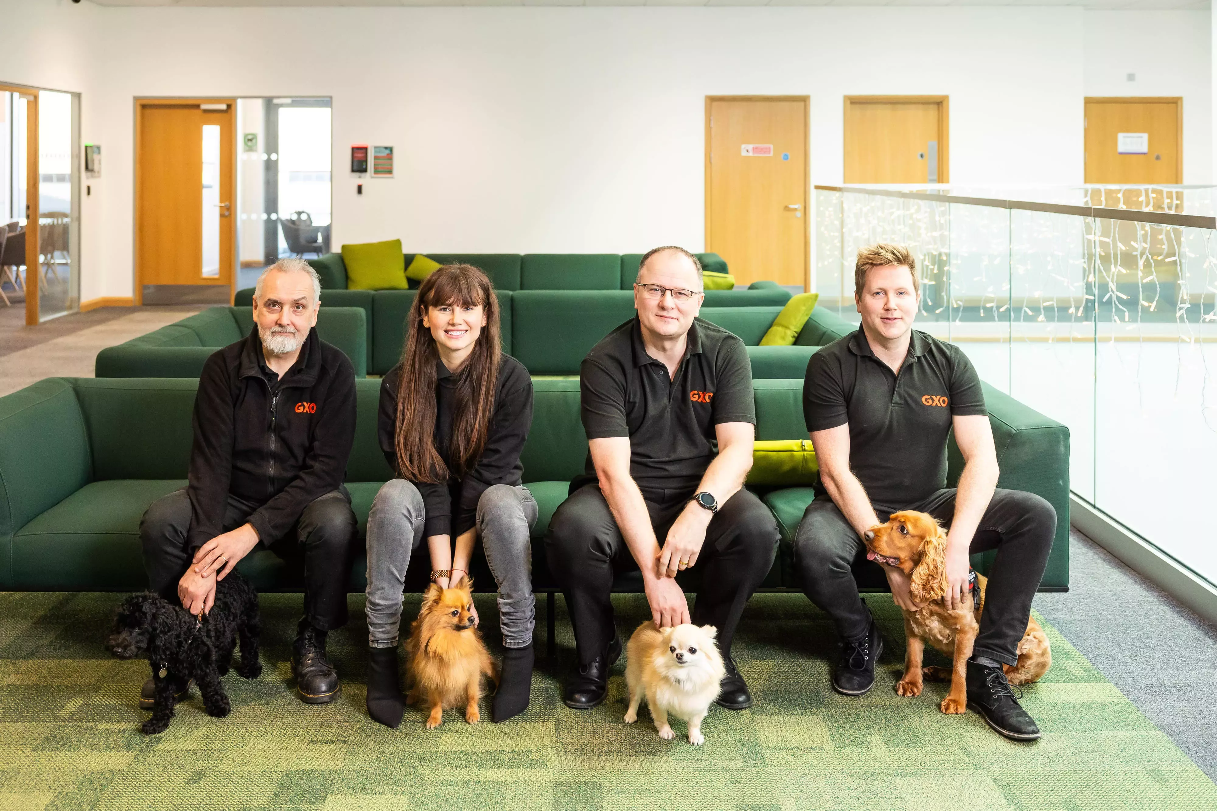 A group of GXO Logistics workers with their dogs beside them, sat on a green sofa inside the office space.