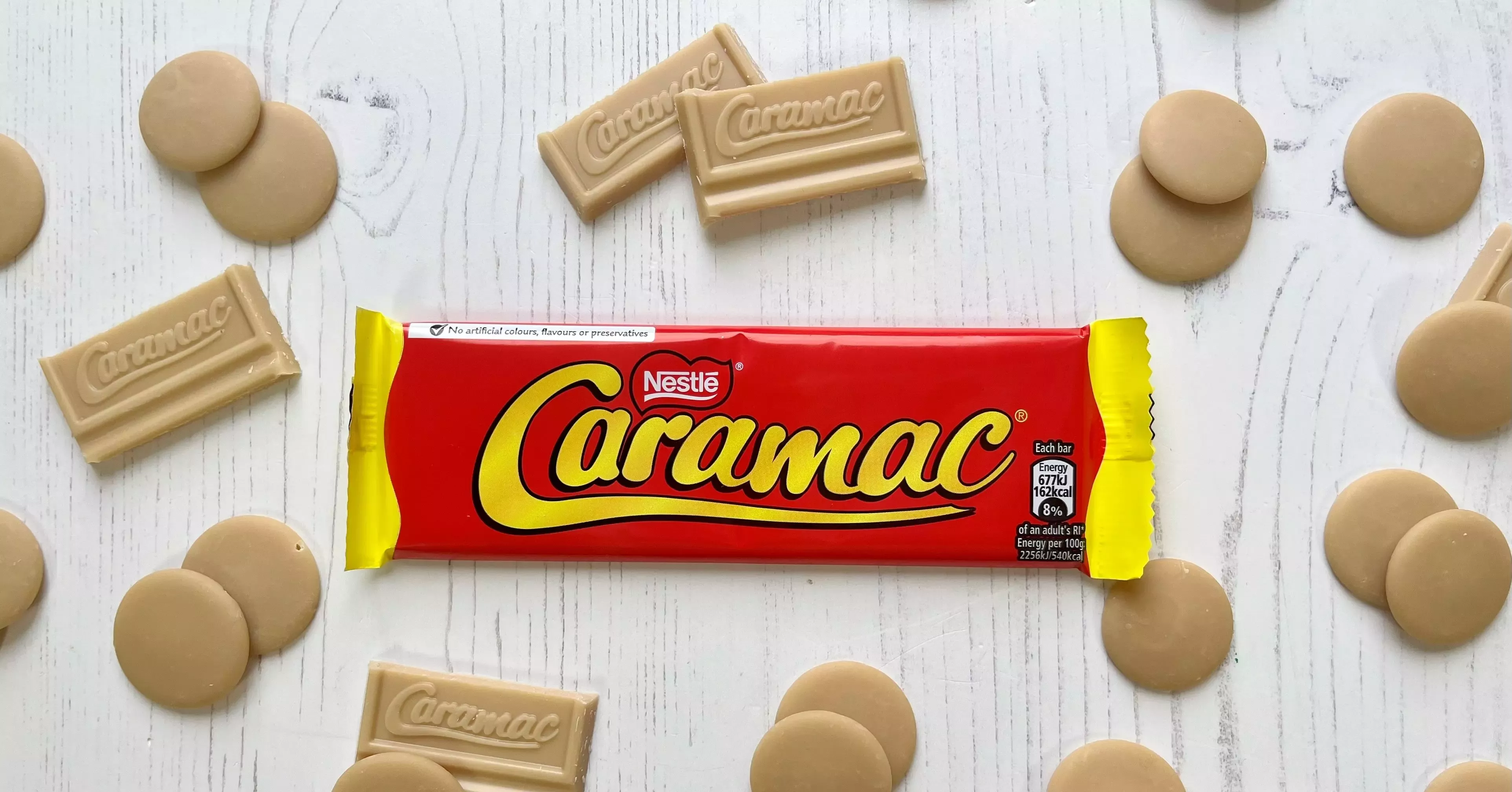 A Caramac bar surrounded by Caramac chocolate pieces on a white wooden background.
