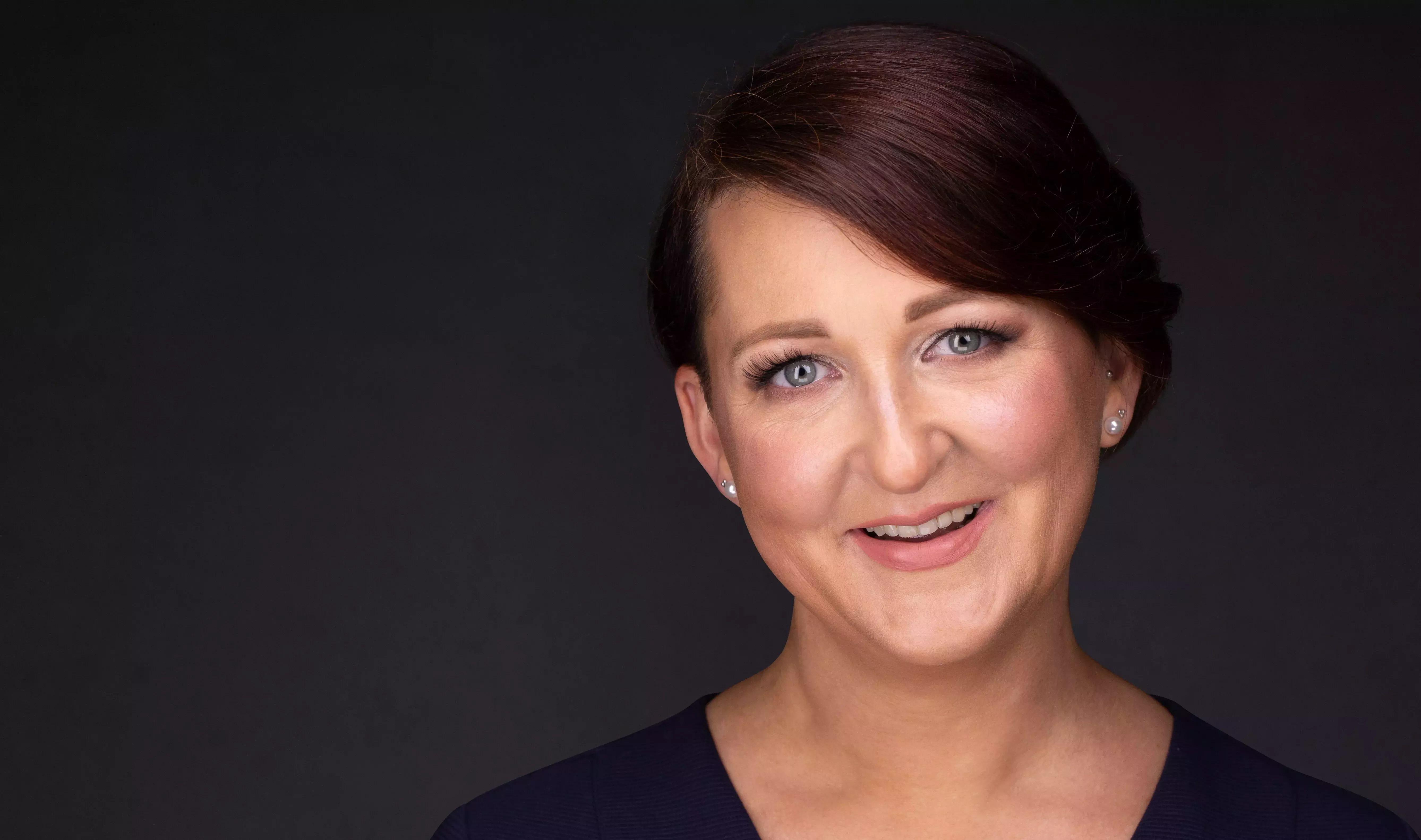 A landscape picture of a smiling woman with short brown hair in a blue top against a grey background.
