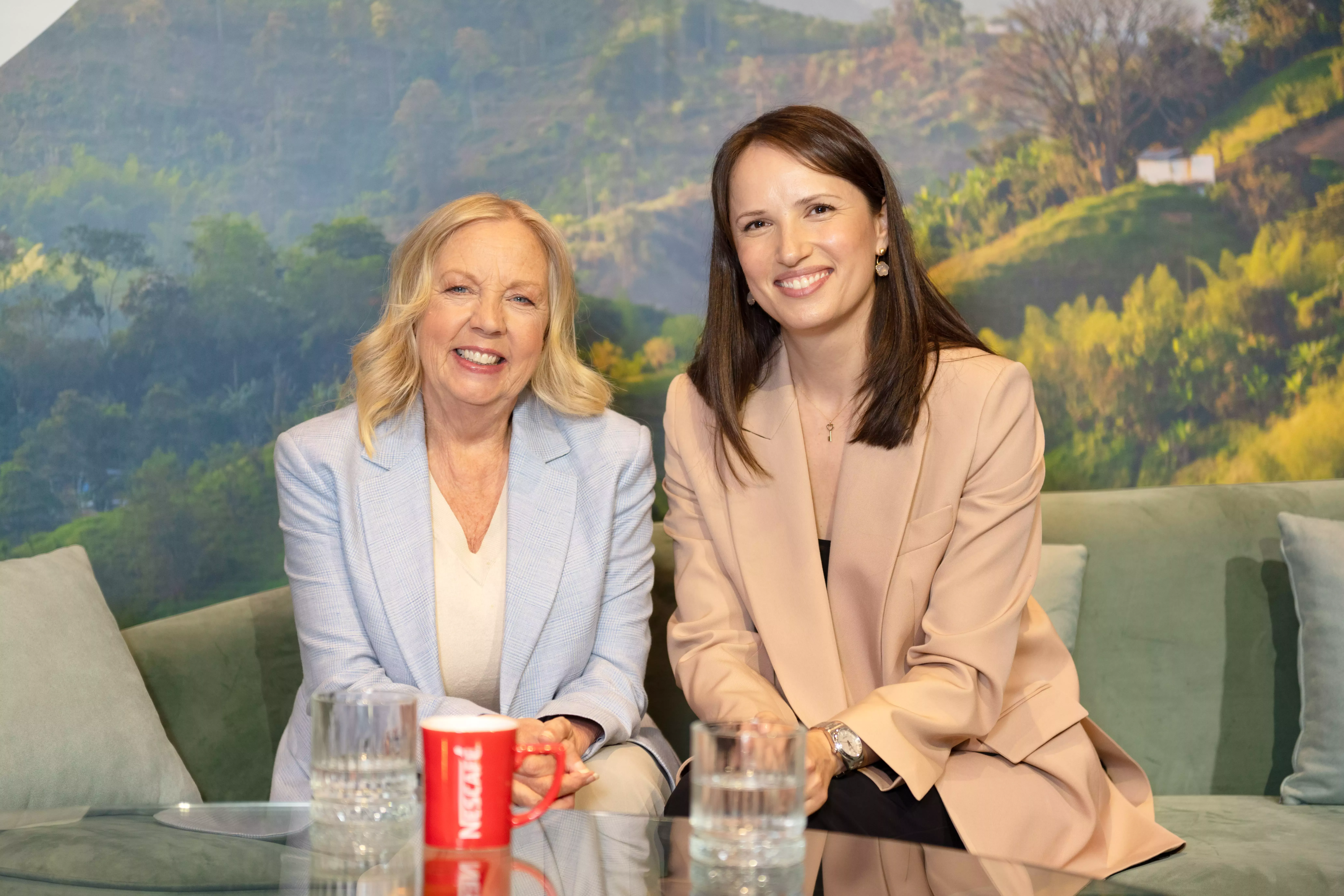 An image of two white females. Deborah Meaden (left) and Katya Simmons (right) sat on a sage green seat, smiling together.