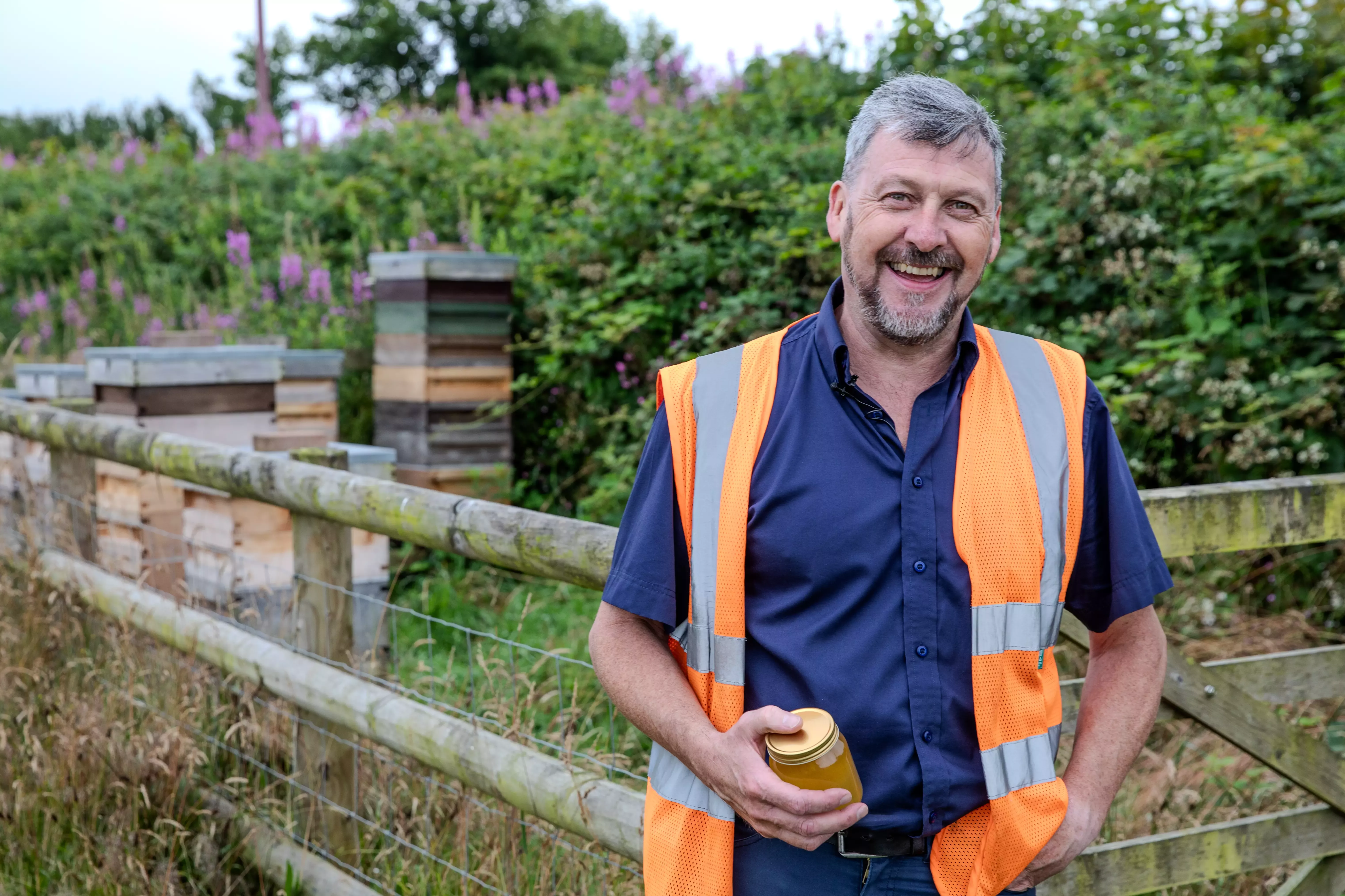 A man in an orange high vis standing in front of beehives and holding a jar of honey. 