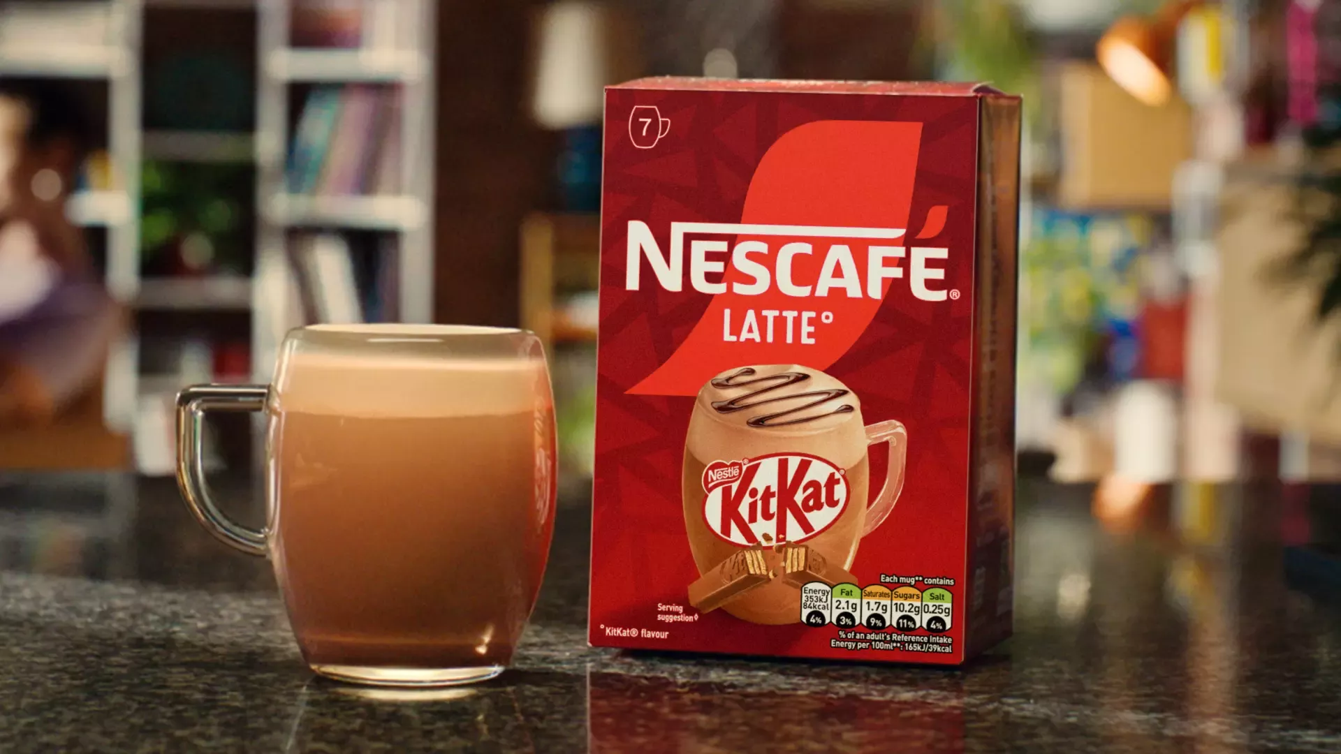 A clear mug filled with Nescafé KitKat coffee next to a dark red product box. The background is warm kitchen setting. 