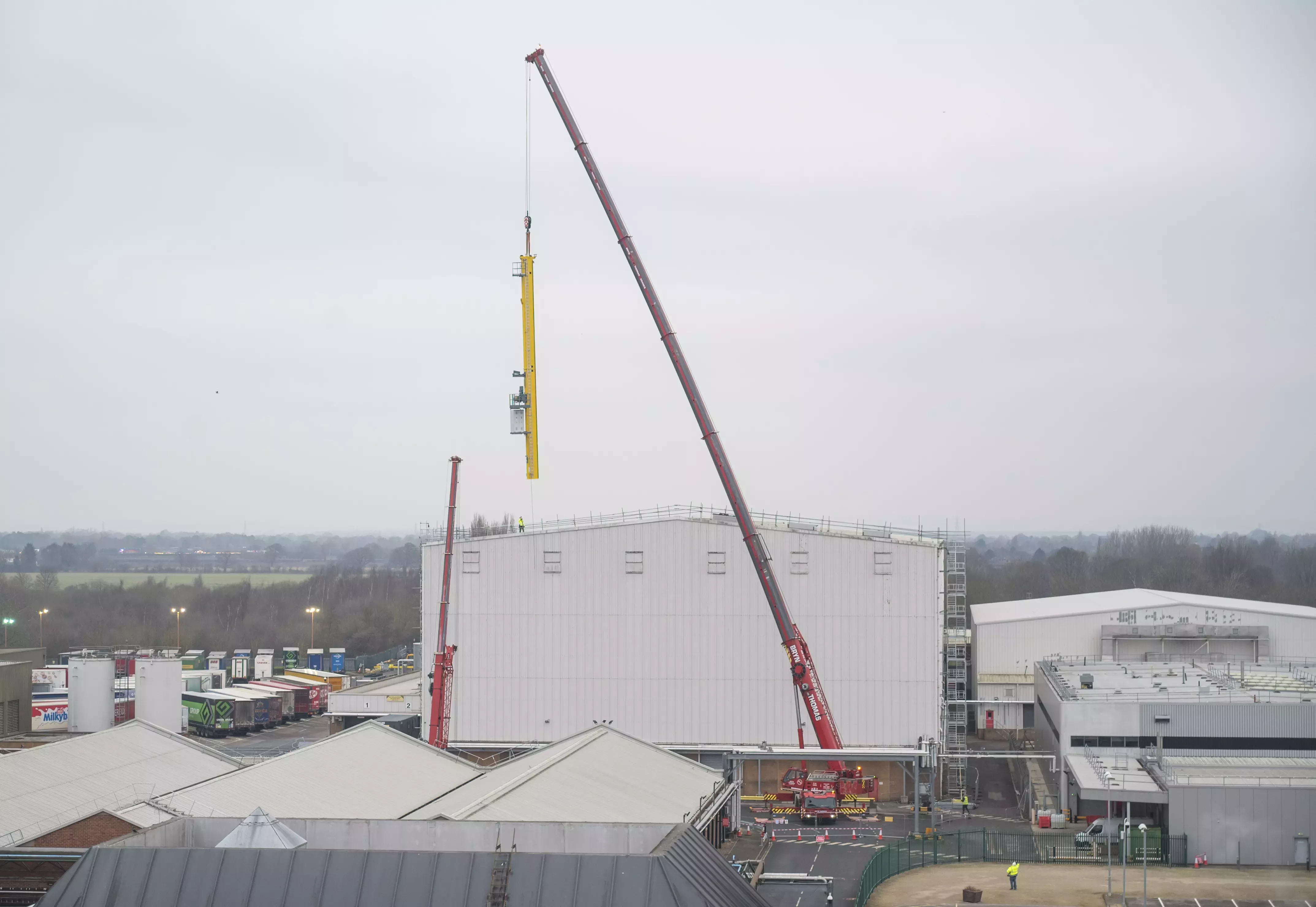 A landscape image of a red 70 metre lifting crane pulling a smaller, yellow crane from a white warehouse roof.