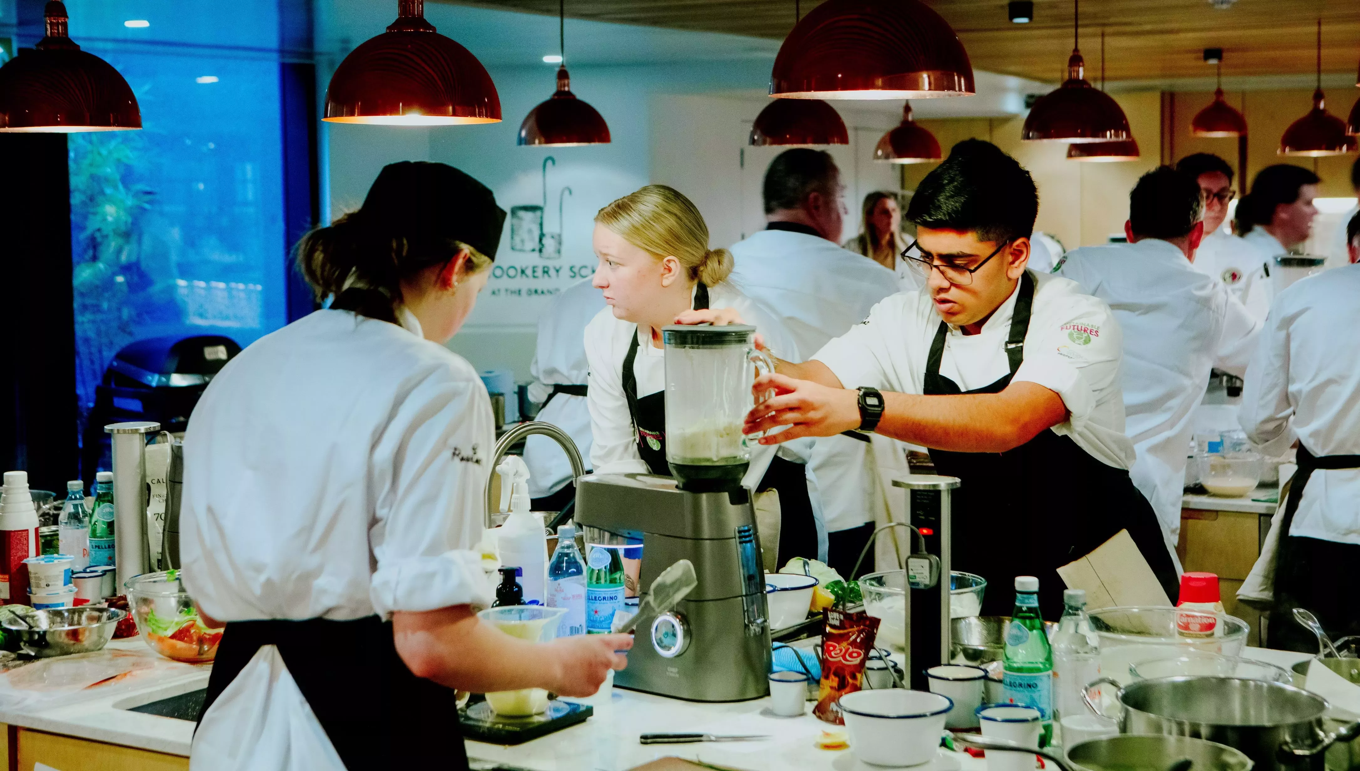 A landscape image of a mixed gender crow of chefs cooking enthusiastically in a modern style kitchen workshop.