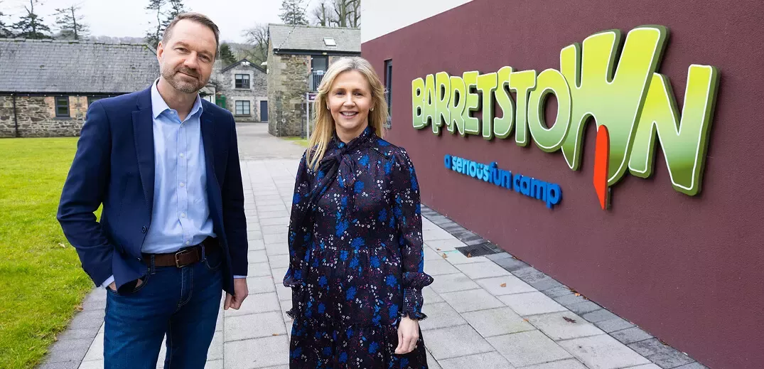 Two individuals stand in front of the colorful Barretstown sign, surrounded by green grass and stone buildings in a serene setting.