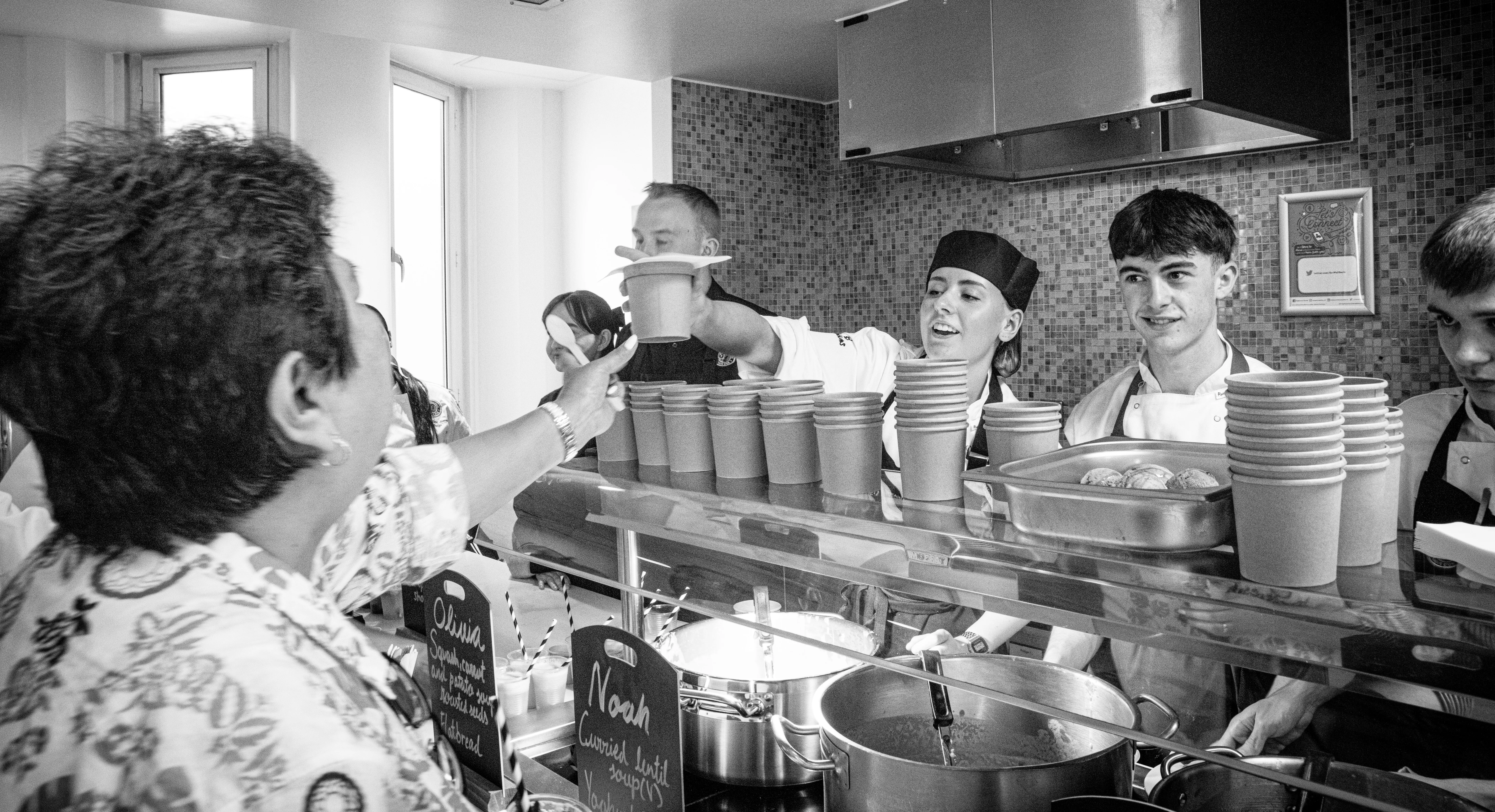 An image of five Toque D'or chefs serving food, one woman is wearing a chefs hat and handing over soup to a customer.