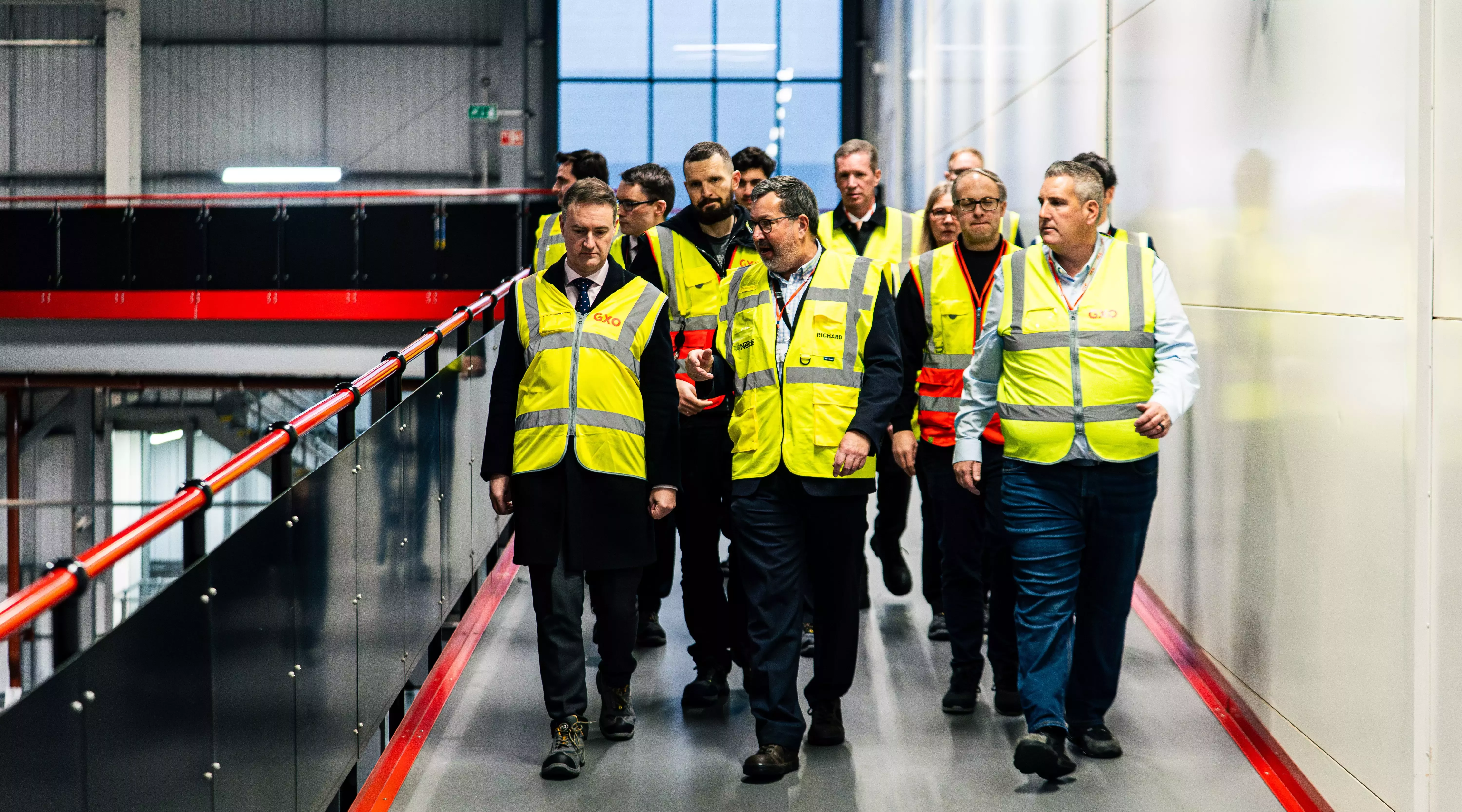 A landscape image of a group of males in yellow hi-visibility jackets walking down the walkway of a distribution centre.
