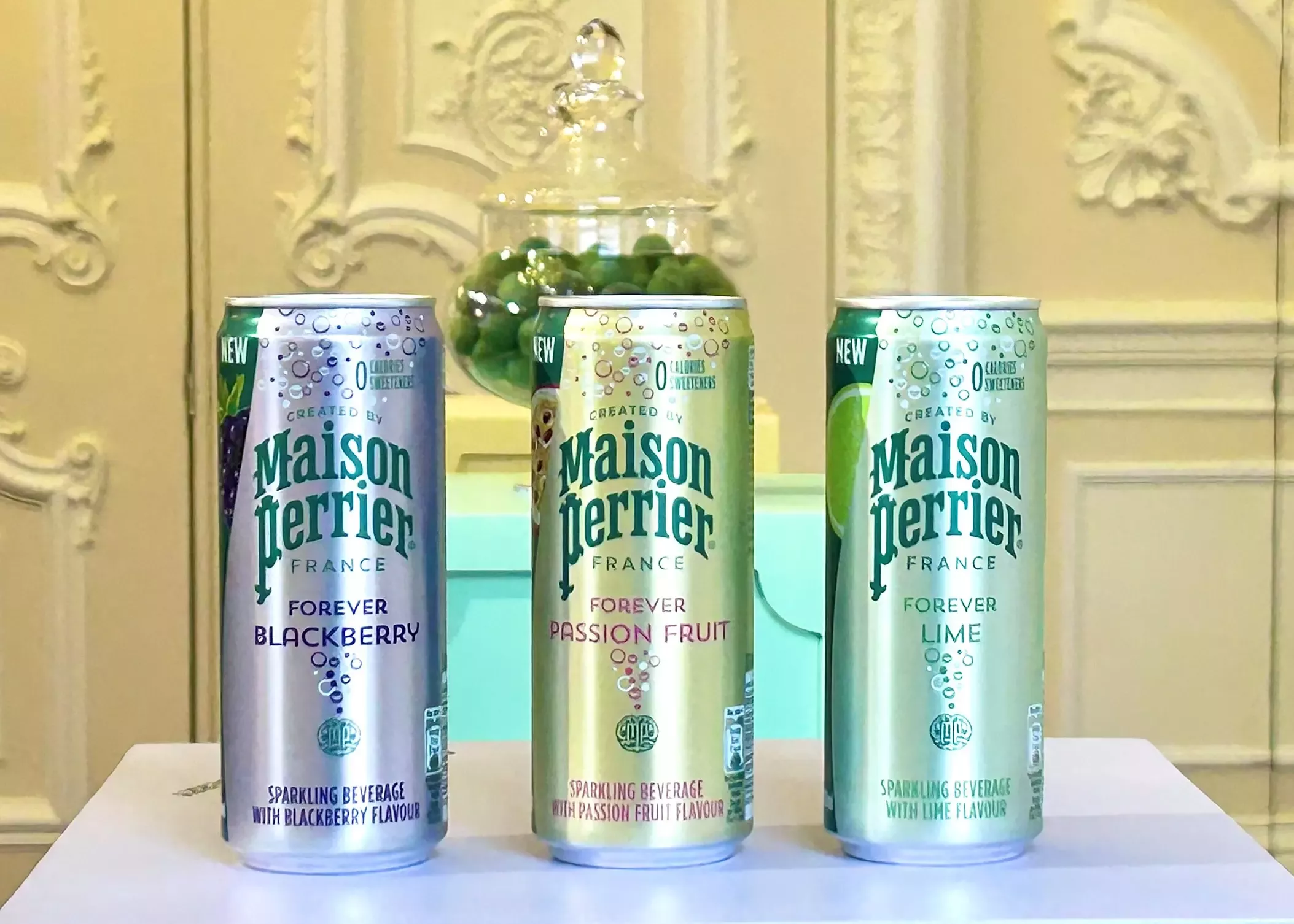 A landscape image of three cans of Maison Perrie, from left to right, the colours of the cans are purple, gold, green. 