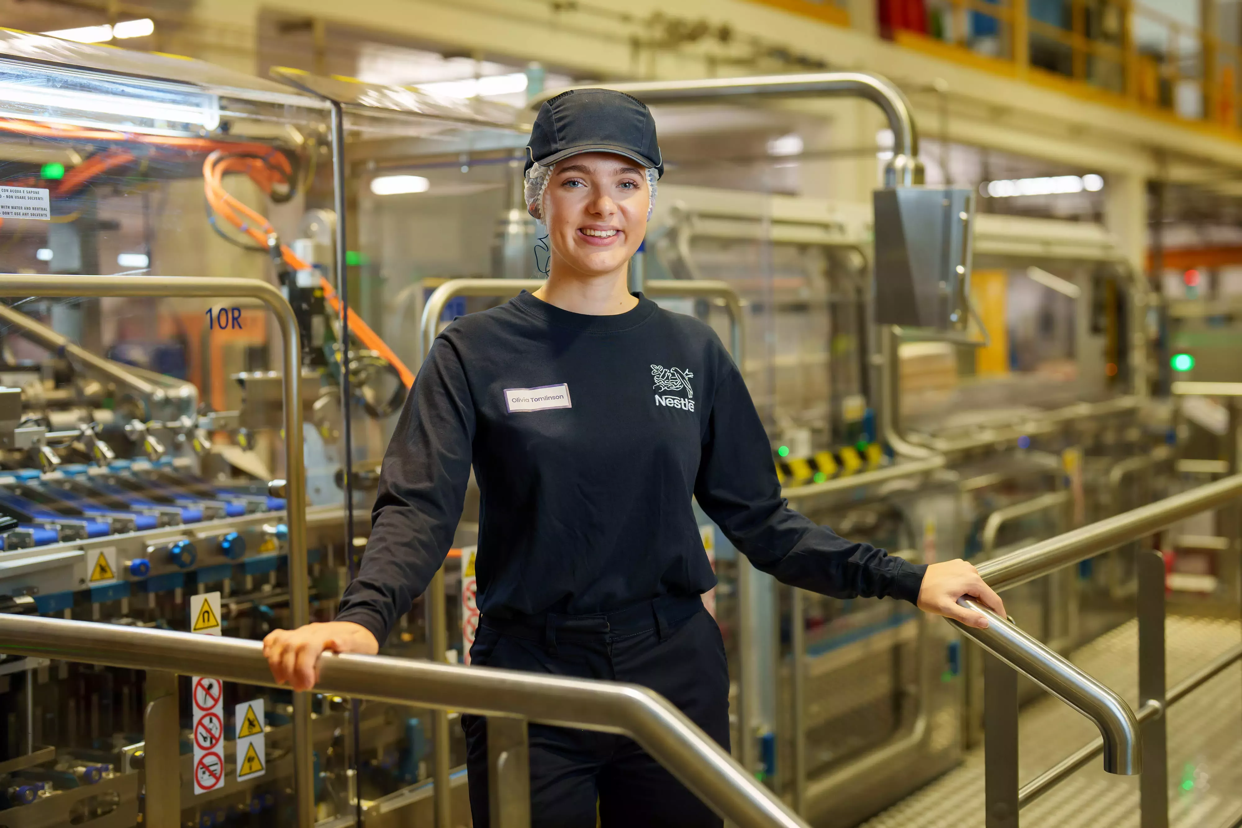 A landscape image of a white young woman, smiling, wearing full factory uniform and a hard hat. 