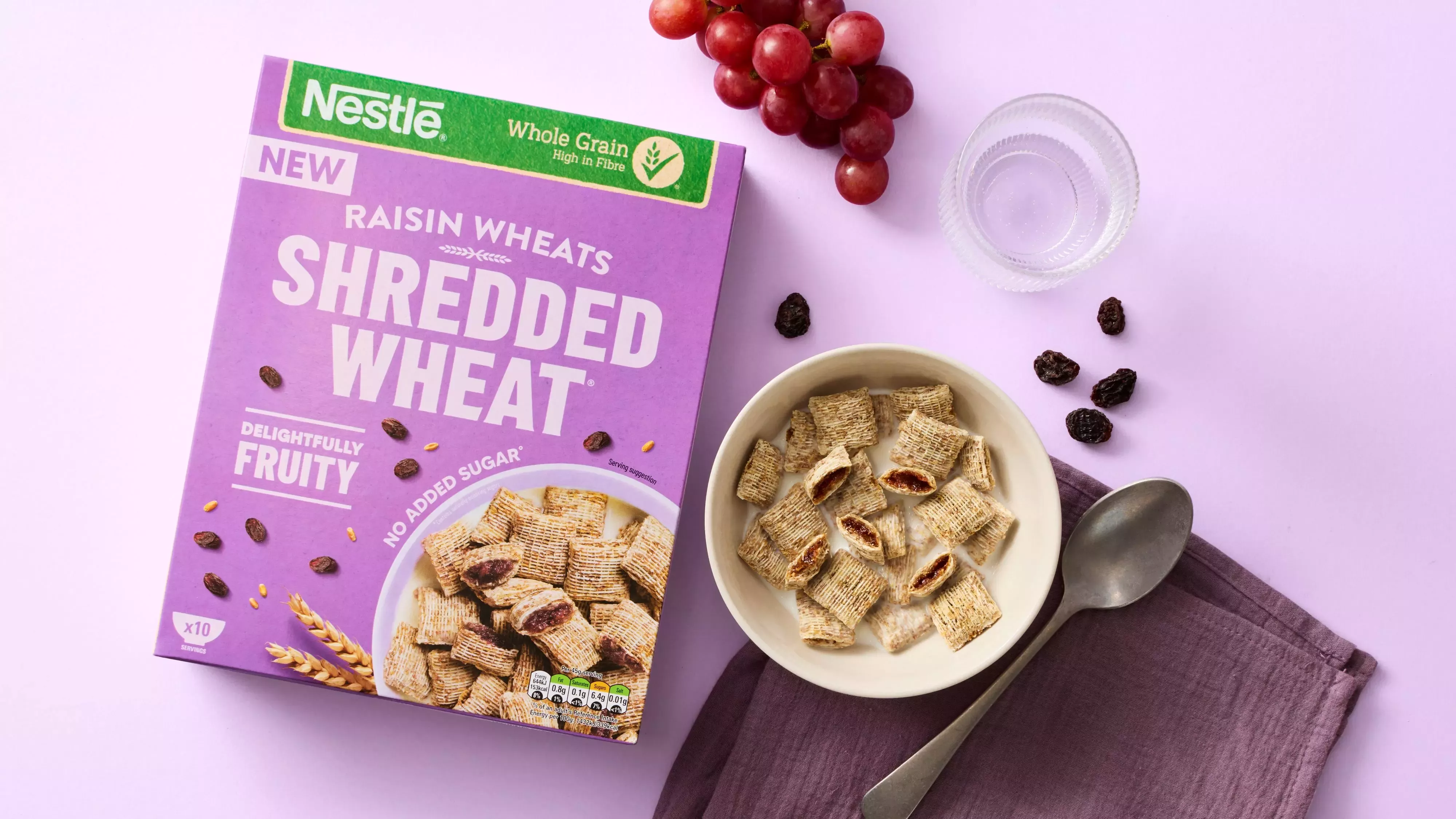Shredded Wheat Raisin Wheats in a bowl next to the box raisins, grapes, a spoon and table cloth