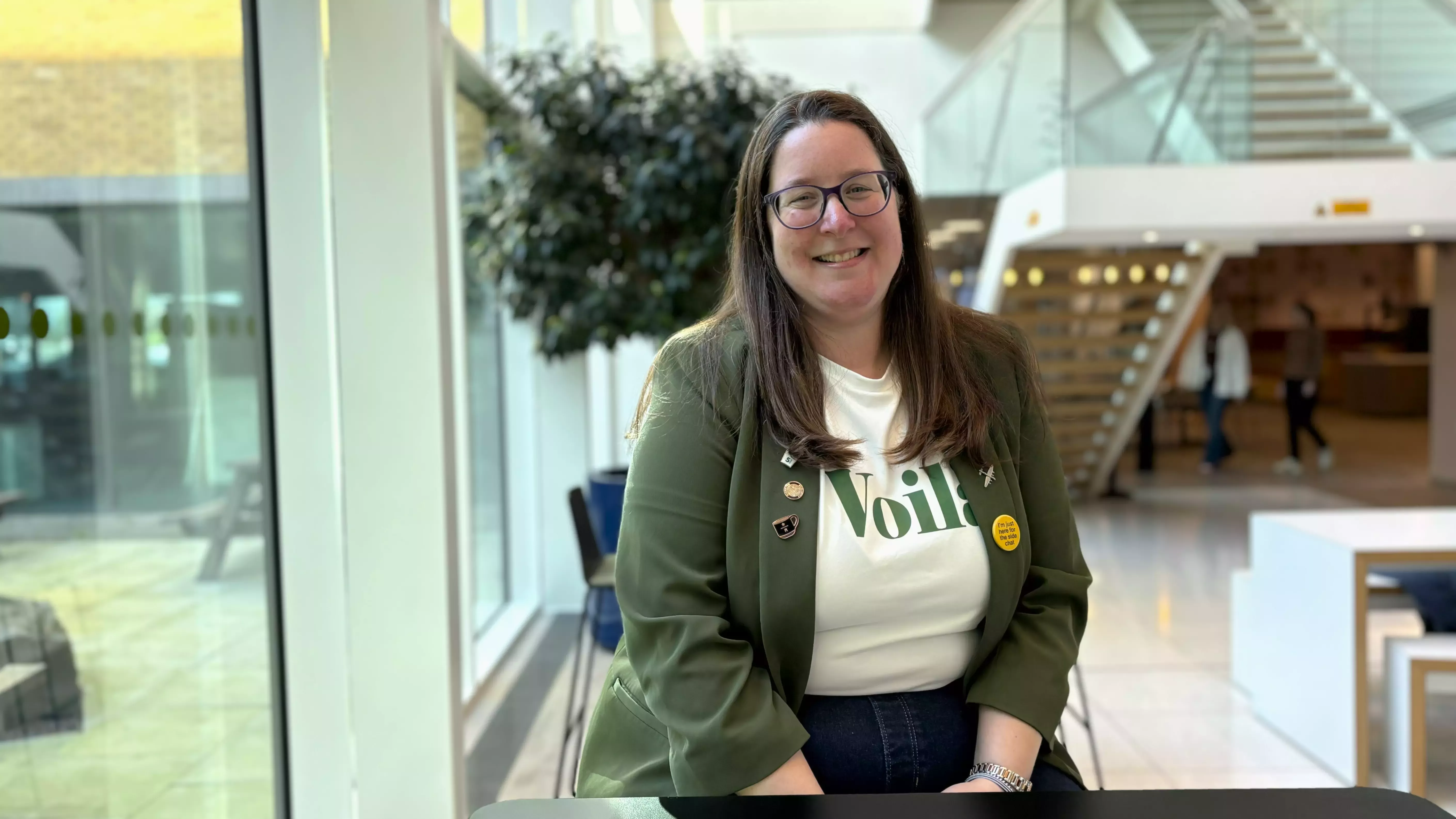 Woman sitting in an office environment wearing a white top and a green blazer.