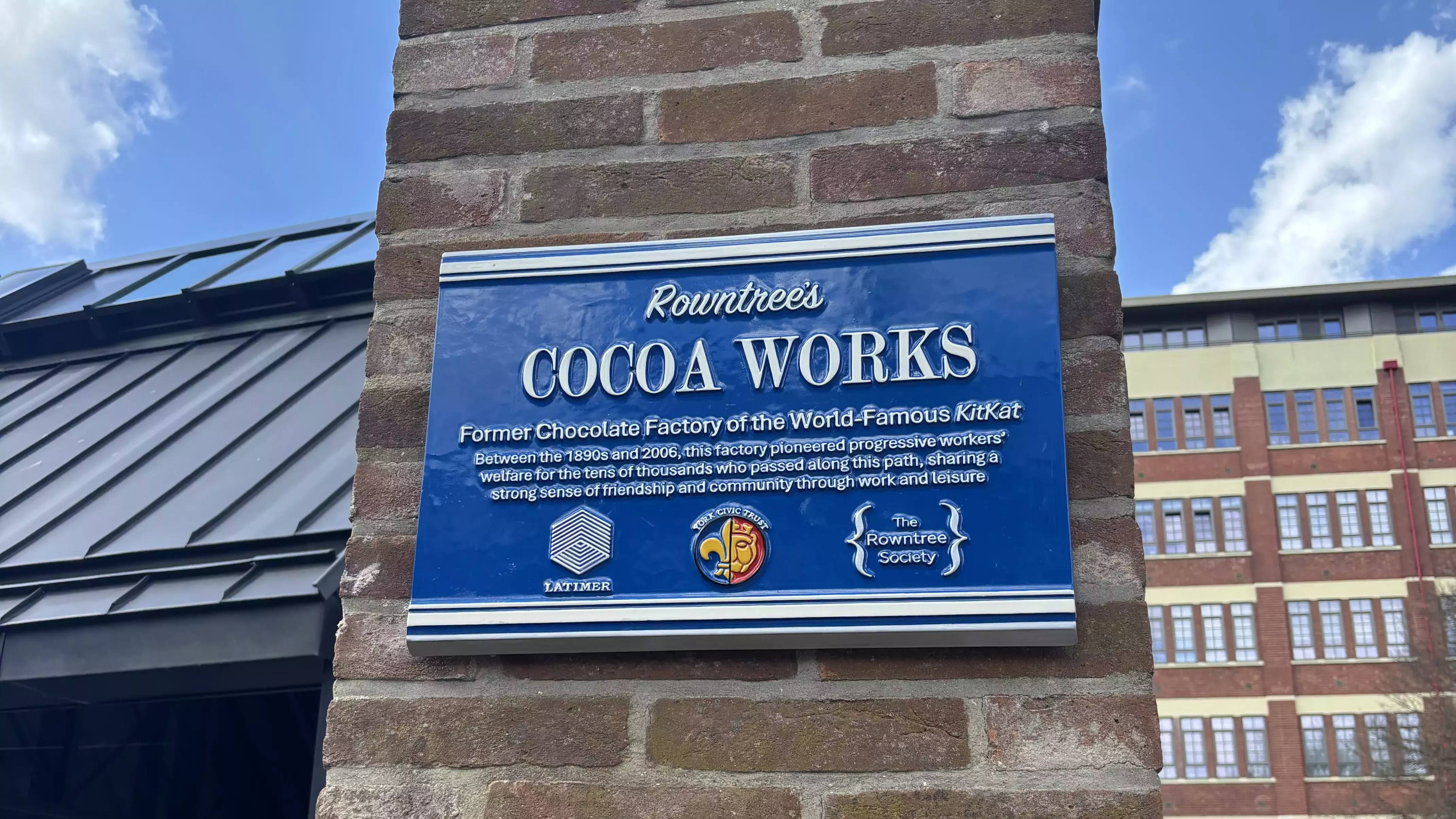 Blue historical plaque on a wall with a building and blue sky in the background.