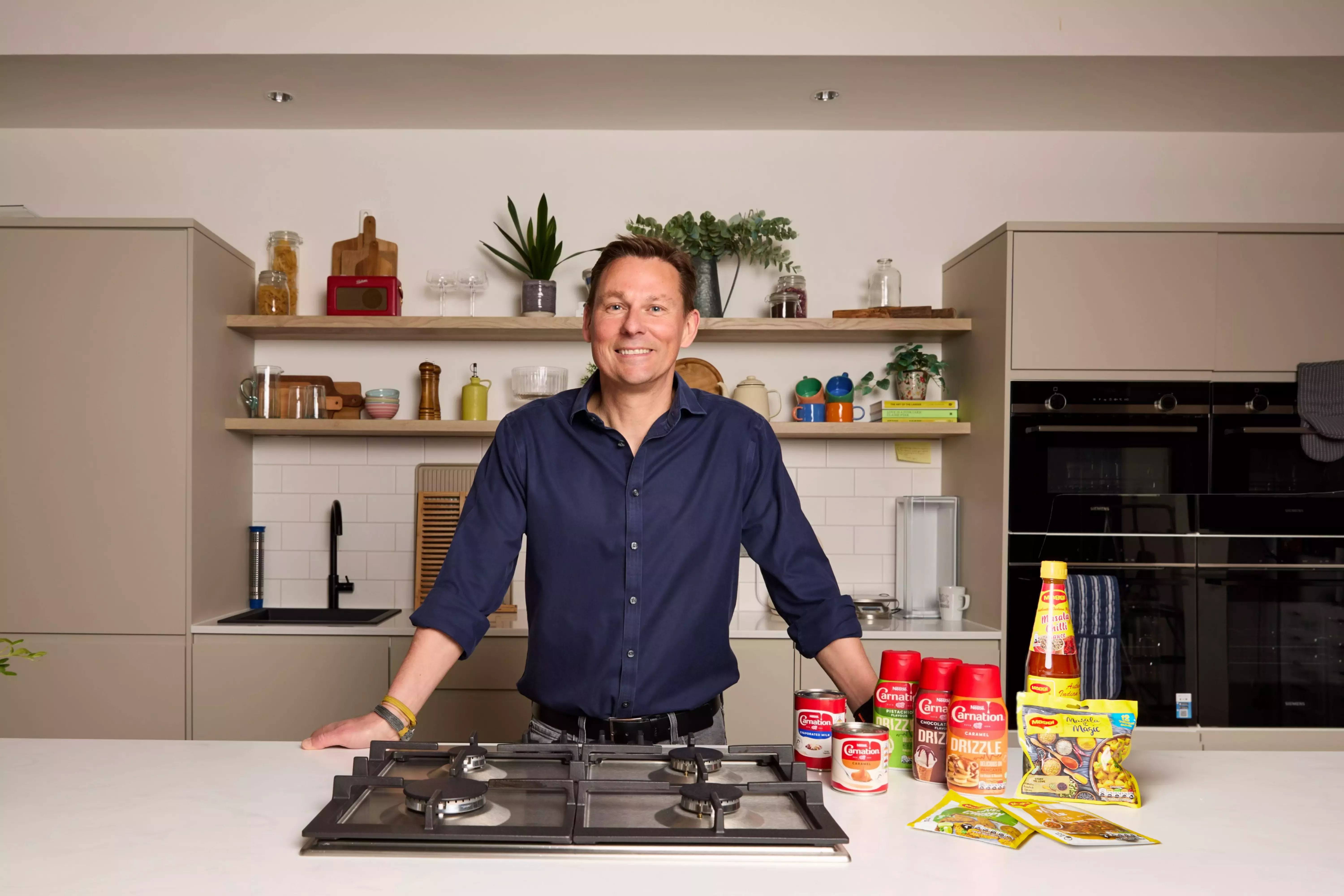 Man standing behind a kitchen counter which has Maggi and Carnation products on it.