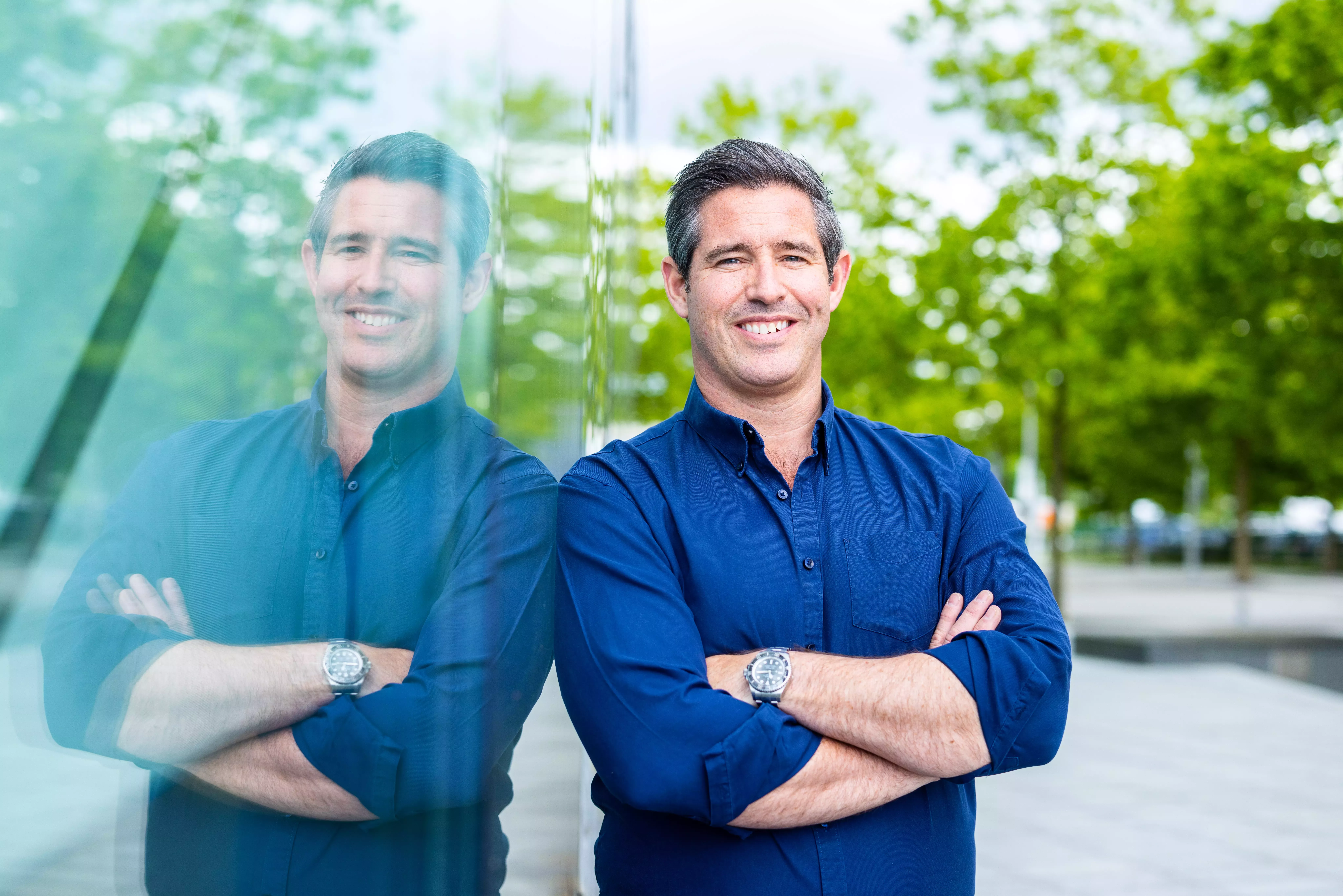 A landscape image of a tall white man in a navy blue shirt, smiling widely and stood against a building window.