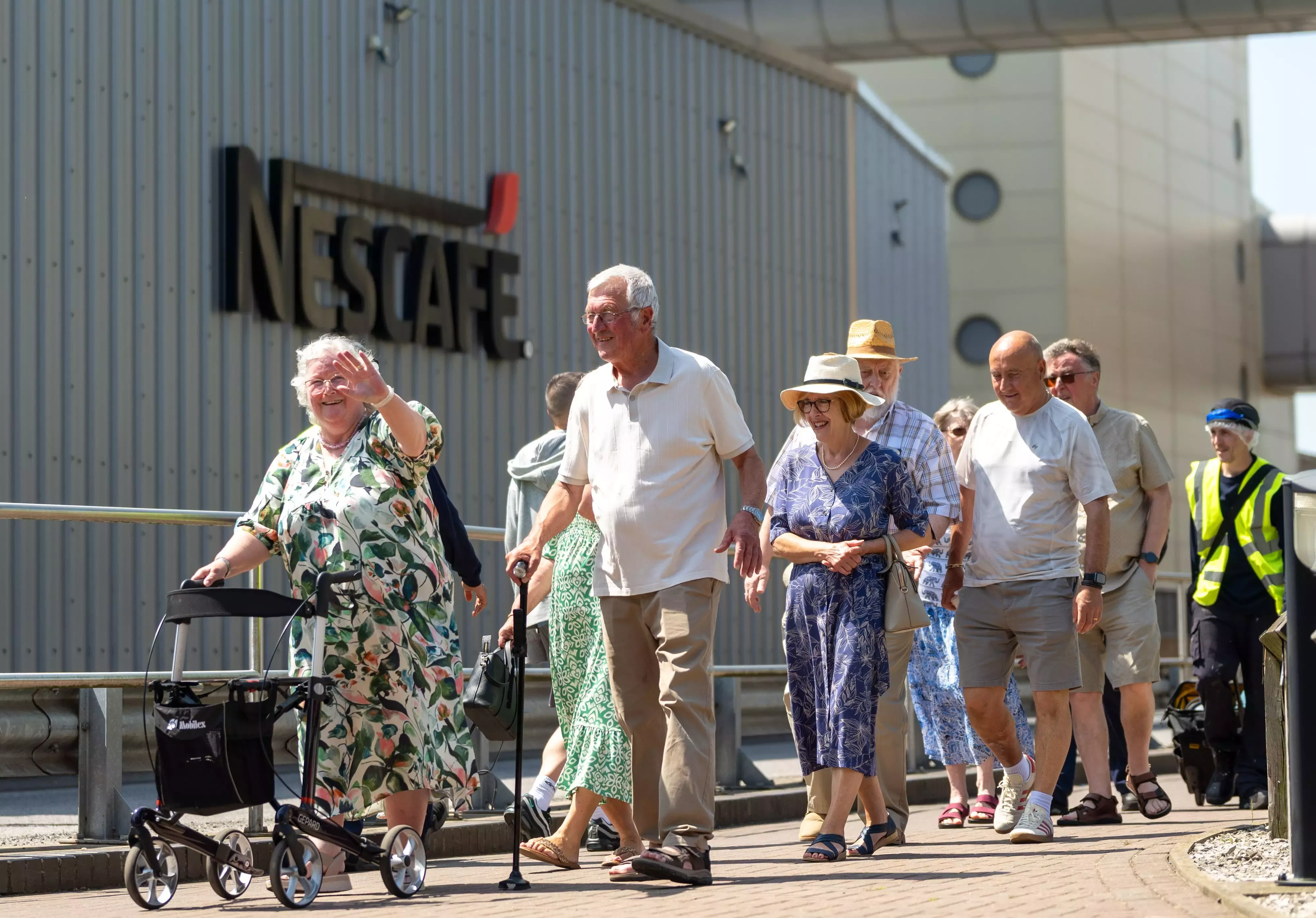 A landscape image of a mixed gender group of local pensioners, smiling and waving outside a Nescafé factory.