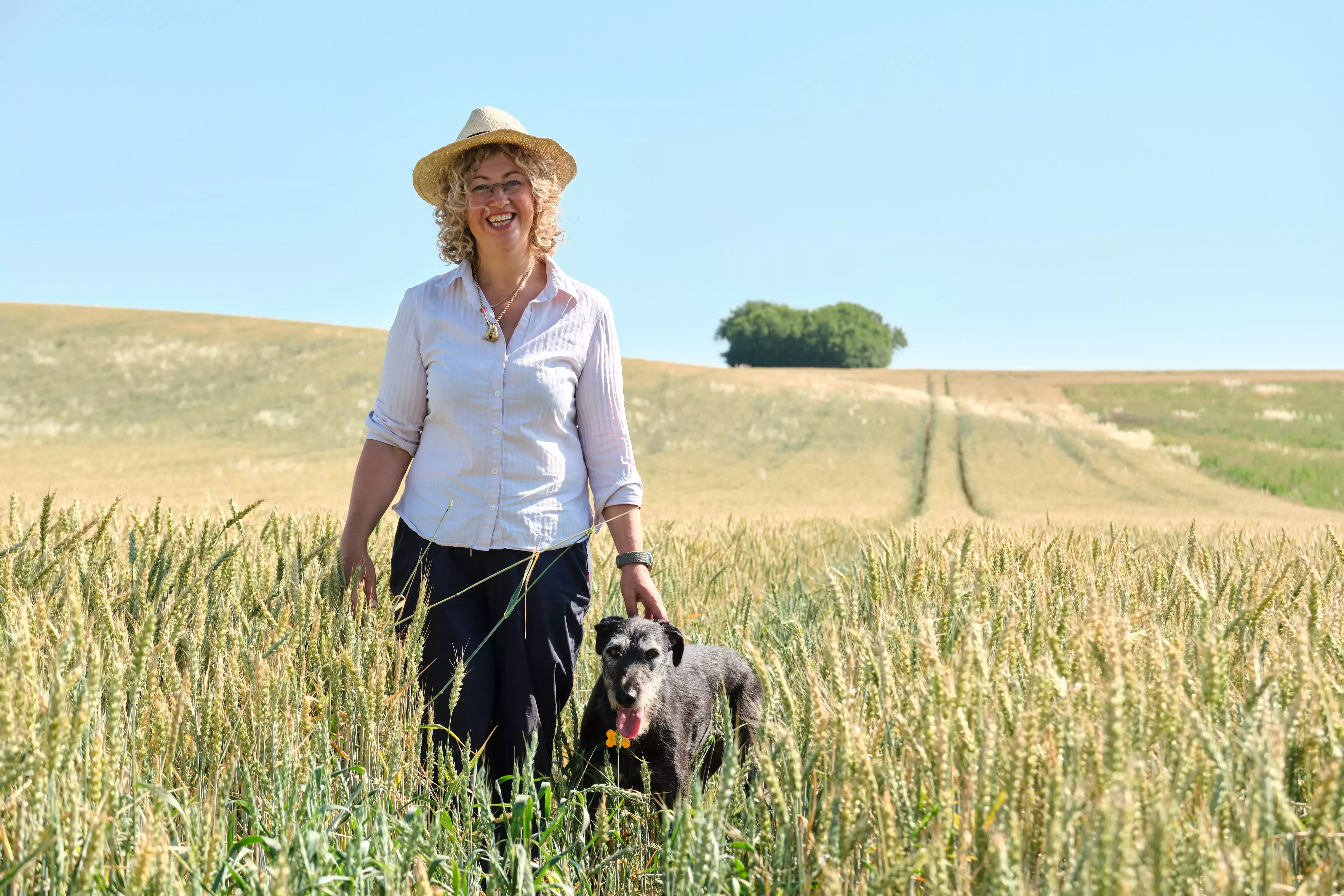 A farmer standing in a wheat field with her grey dog by her side.