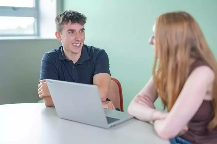 Two people seated at a table discussing work with a laptop open between them.