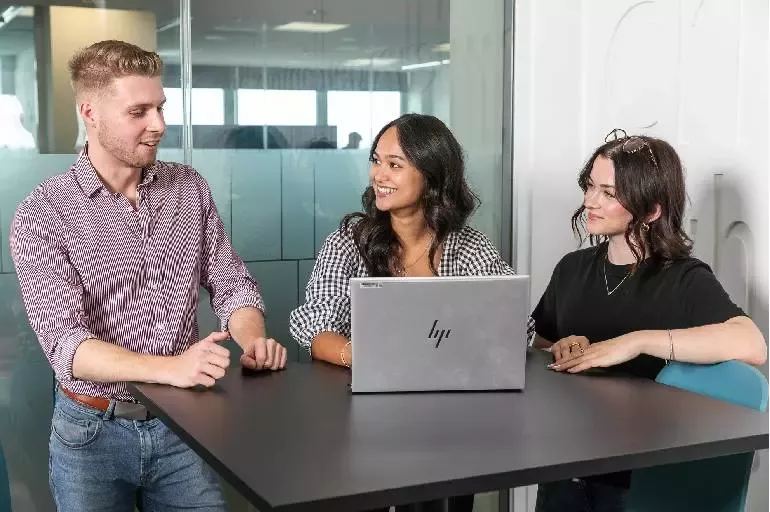 Three people seated at a meeting table discussing work while using a laptop in an office environment.