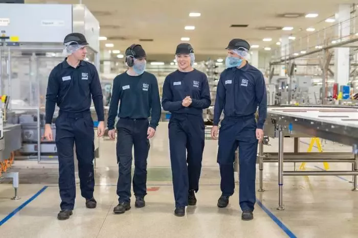  Four people in factory workwear walking through a manufacturing facility alongside a production line.