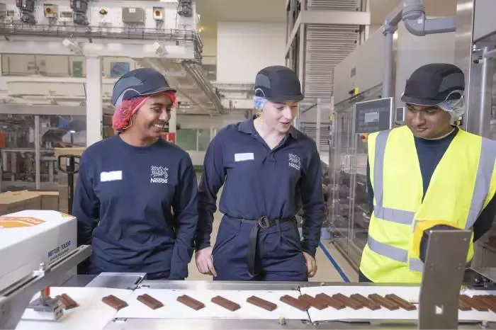 Three apprentices wearing workplace safety equipment standing beside a production line in a food manufacturing facility.