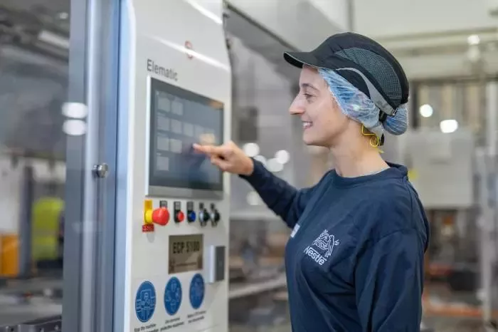Apprentice operating a touchscreen control panel on manufacturing equipment in a food production facility while wearing workplace safety gear.