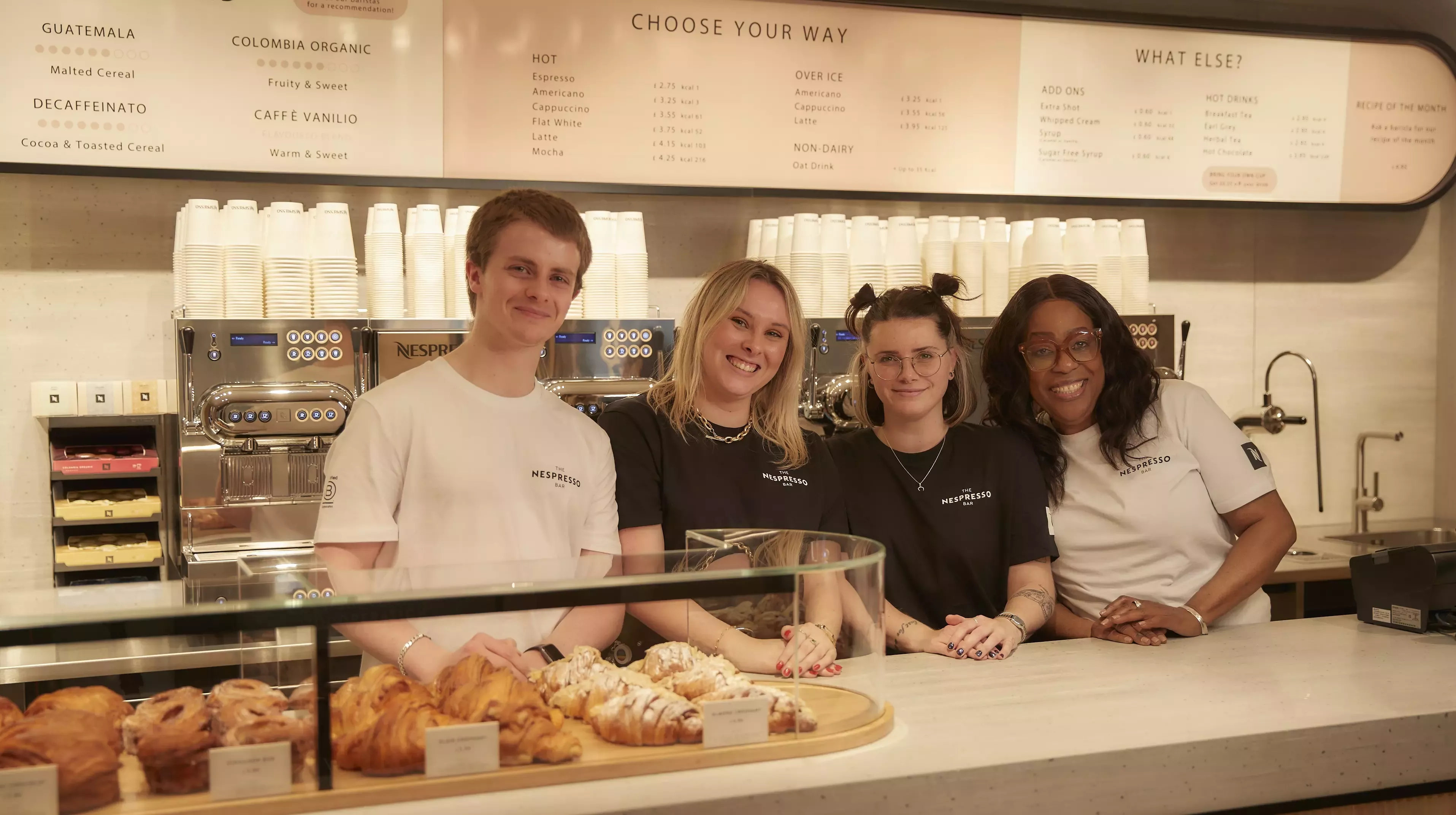 A cafe counter top with coffee machines and pastries on top, standing behind and smiling is one man and three women.
