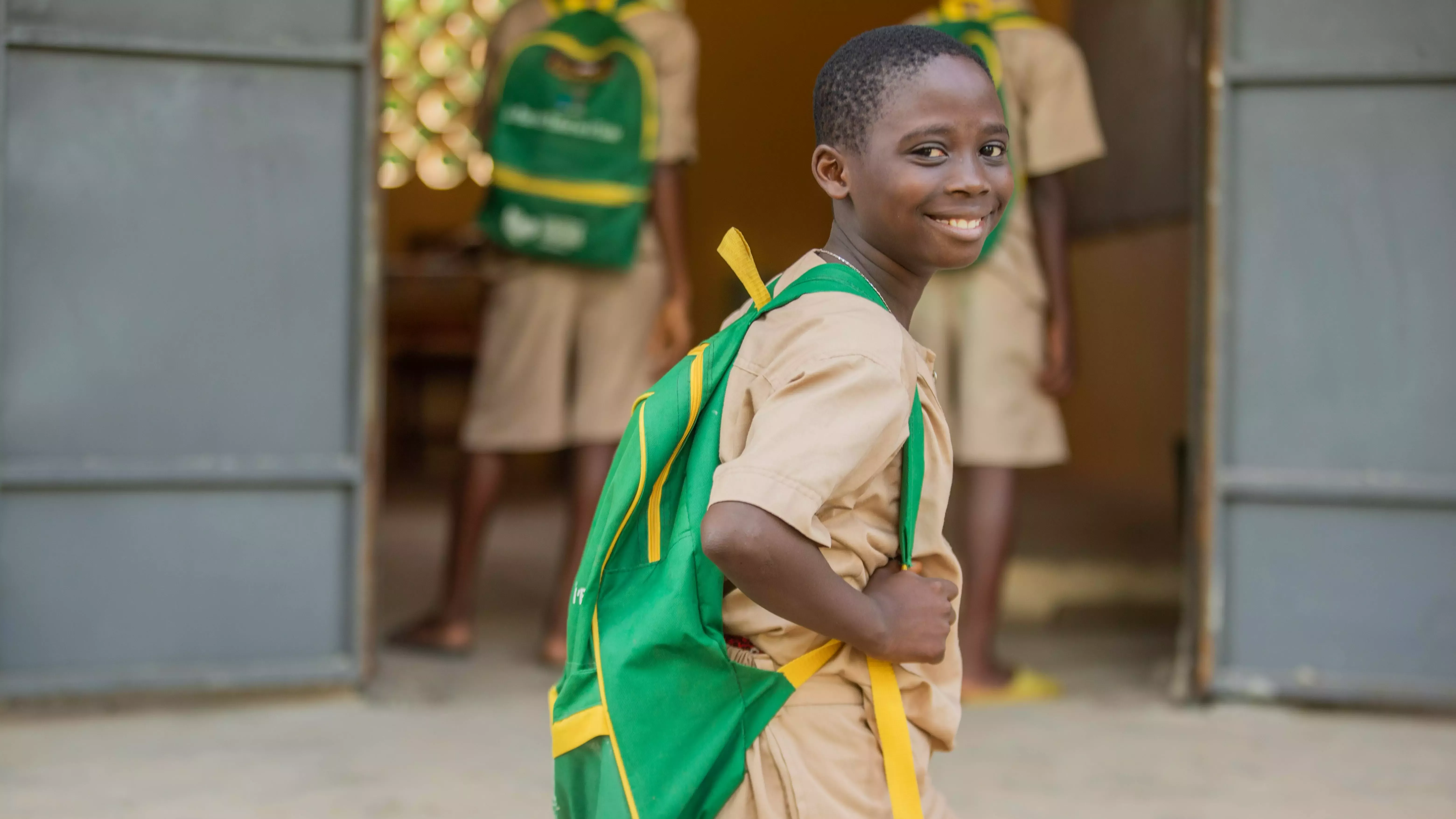 Young boy, smiling, and wearing a brown t-shirt and green backpack.